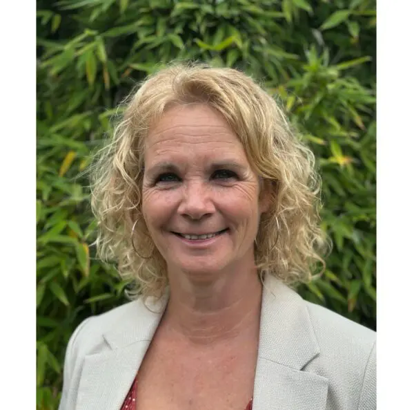 Portretfoto van een vrouw met blond krullen halflang haar. Ze draagt oorbellen. Haar ogen zijn donker. Ze draagt een grijs colbert. De achtergrond bestaat uit groene bladeren.