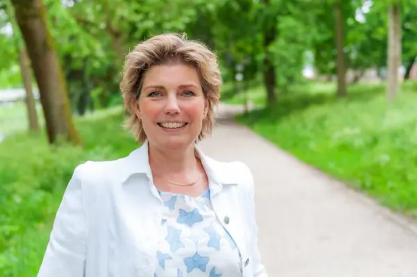 Foto van een vrouw die breeduit lacht, ze heeft kort blond haar. Haar wit blauwe bloemetjes blouse en haar witte jasje steken af tegen het groen van de achtergrond.