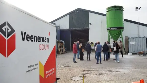 Een groep deelnemers op het terrein van Veeneman. Ze staan voor een schuur met een puntdak en een groene watertoren. Op de voorgrond staat een busje met het logo van het bedrijf.