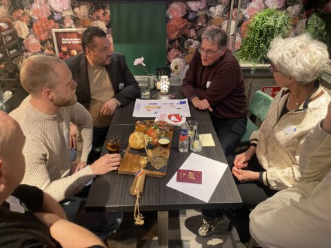 Een groep van zes mensen zitten om een tafel heen met werkdocumenten, drankjes en broodjes.