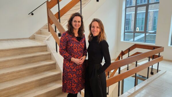 Spinners Eefje Remijn en Anne-Louise Bergkamp staan in een trappenhuis met grote ramen op de achtergrond. Eefje staat links, ze heeft bruin haar en een rode jurk aan. Anne-Louise staat rechts, ze heeft blond haar en een zwarte jurk aan.